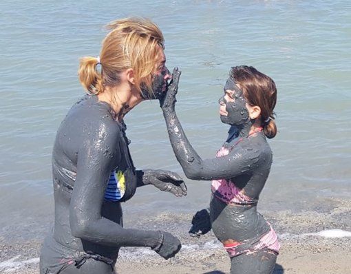 Woman and child covered in mud, applying it to each other's faces by a body of water.