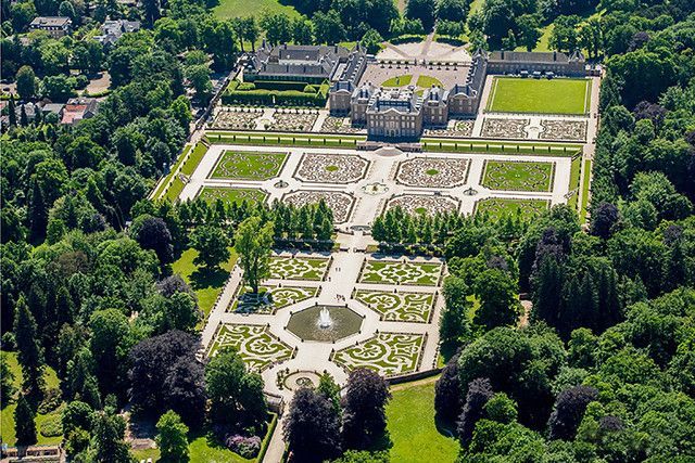 Aerial view of a grand European palace with symmetrical gardens, lush green lawns and surrounding trees.
