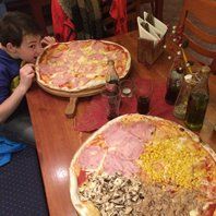Boy leans towards large pizza on a wooden board. Another giant pizza, drinks, and table setting.