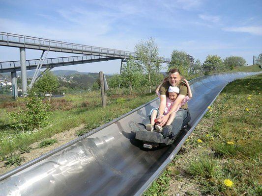 Man and child ride a metal slide down a grassy hill, with a raised walkway structure in the background.
