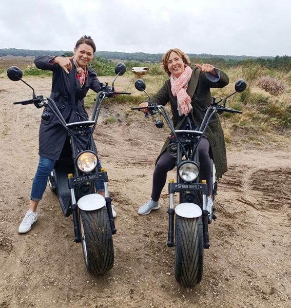 Two women on white electric scooters, thumbs-up gesture, outdoor setting.