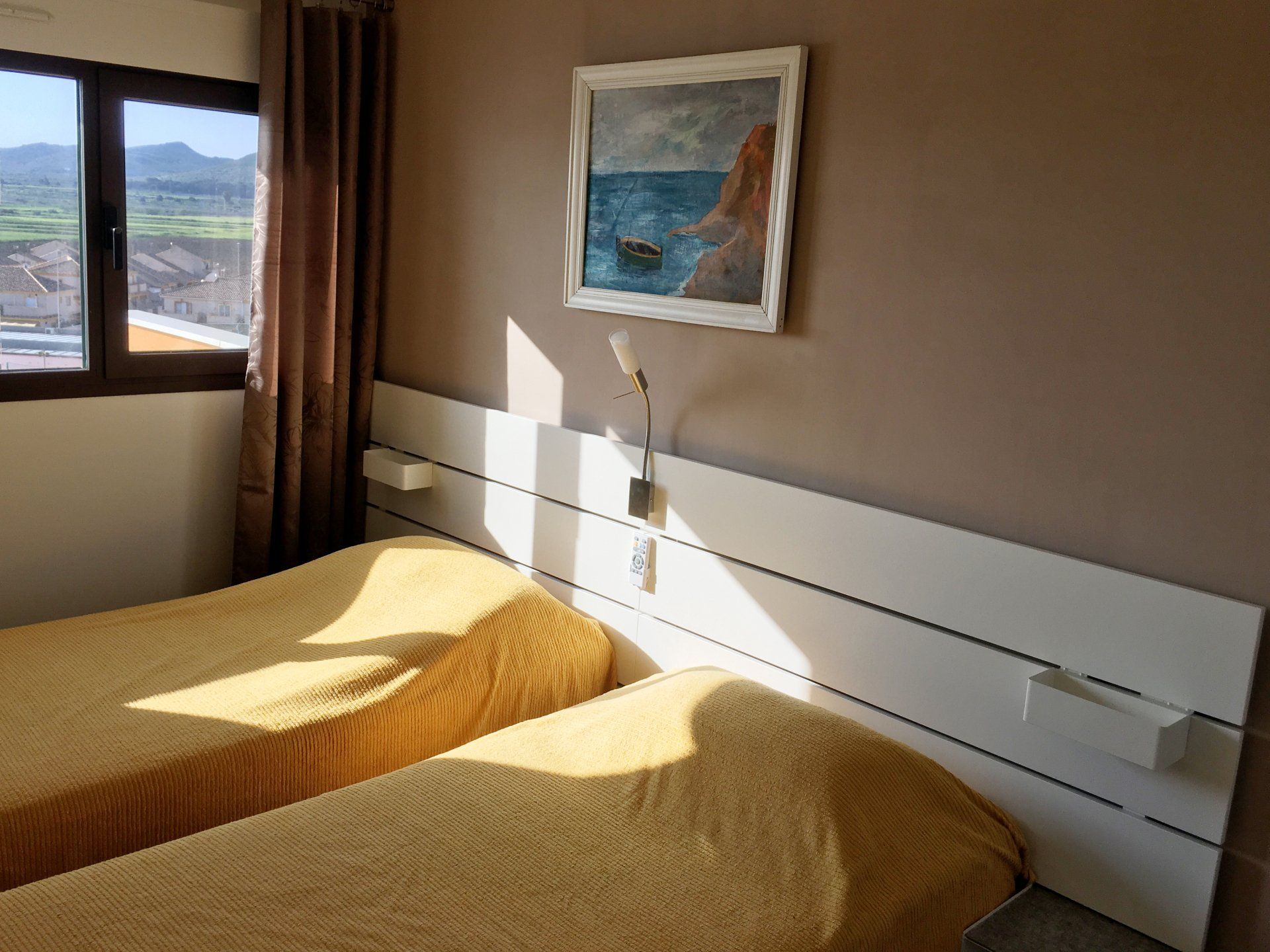 Bedroom with two twin beds, tan walls, window with brown curtains, and a seascape painting.