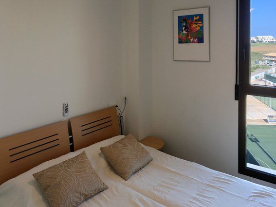 Bedroom with a bed, pillows, nightstand, framed artwork, and window overlooking a cityscape.