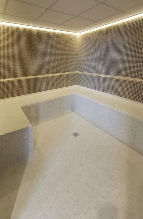 Spa room with light-colored tile walls, a built-in bench, and a drain. Warm lighting along the ceiling.