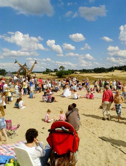 People gathered in a sandy area under a blue sky, with giraffe sculptures in the background.