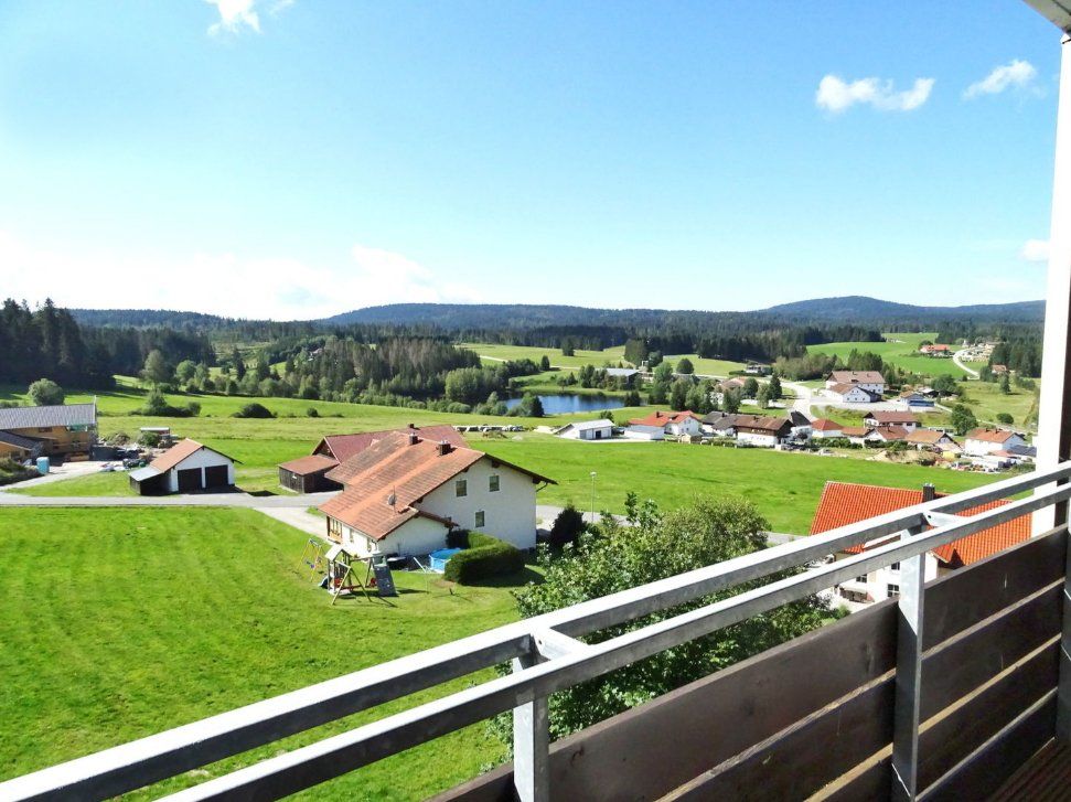 View from a balcony overlooking a green field with buildings and a forest on a sunny day.