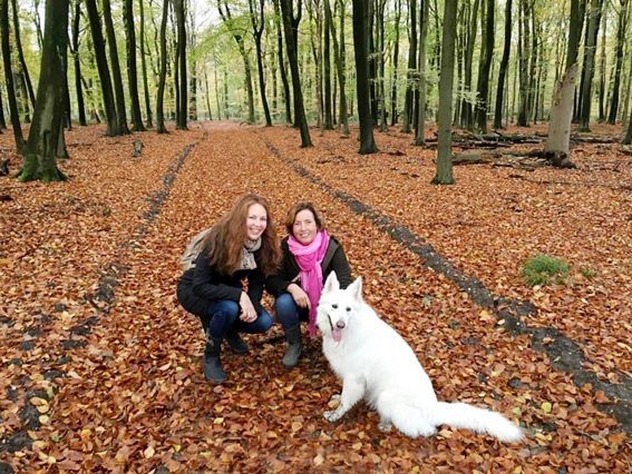 Two people and a white dog crouch on a leaf-covered path in a forest.