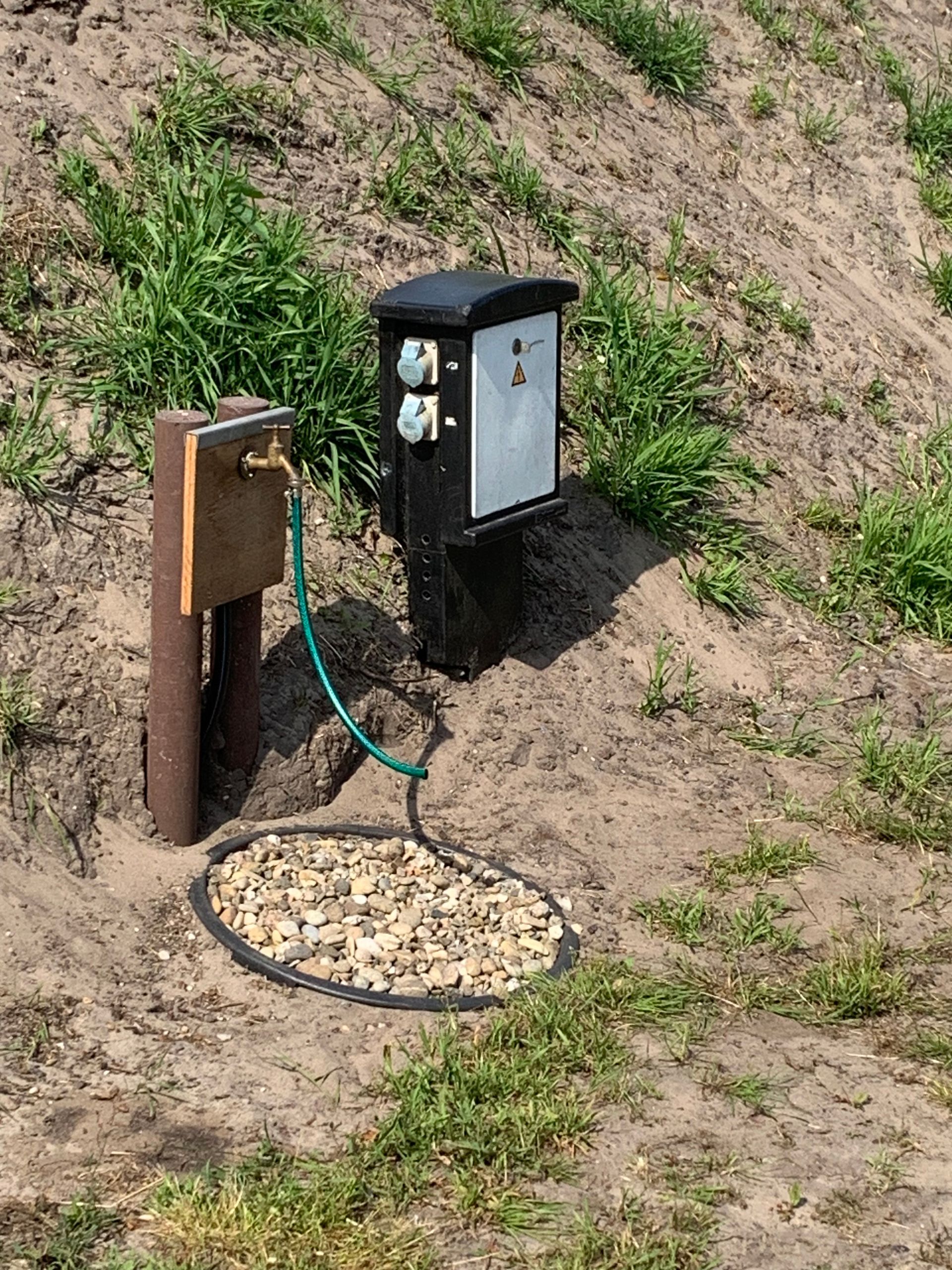 Electrical and water hookups at a campsite with a green hose, black box, and brown pipe set in the dirt.