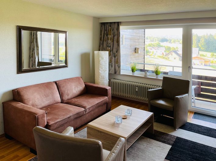 Living room with brown sofa, two chairs, coffee table, large window overlooking a balcony.