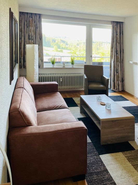Living room with brown sofa, armchair, and coffee table; large window with view.