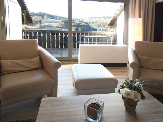 Cozy living room with two beige armchairs, ottoman, and balcony overlooking mountains.