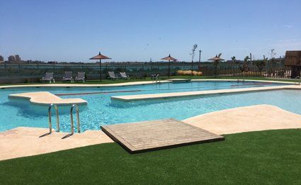 Swimming pool with blue water, surrounded by concrete and green artificial grass, under a clear sky.