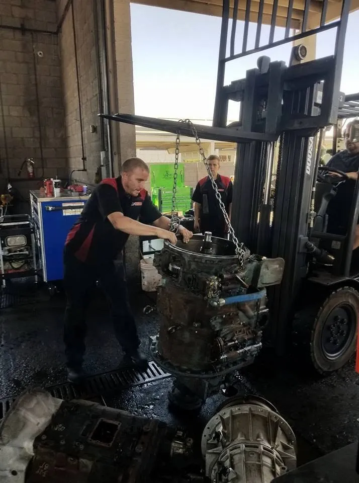 Mechanic using a forklift to move a heavy engine in a repair shop. Another man observes.