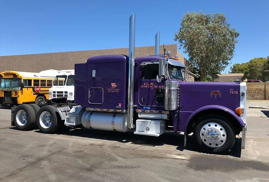 Purple semi-truck with silver accents parked outside. A school bus and a white van are nearby under a sunny sky.