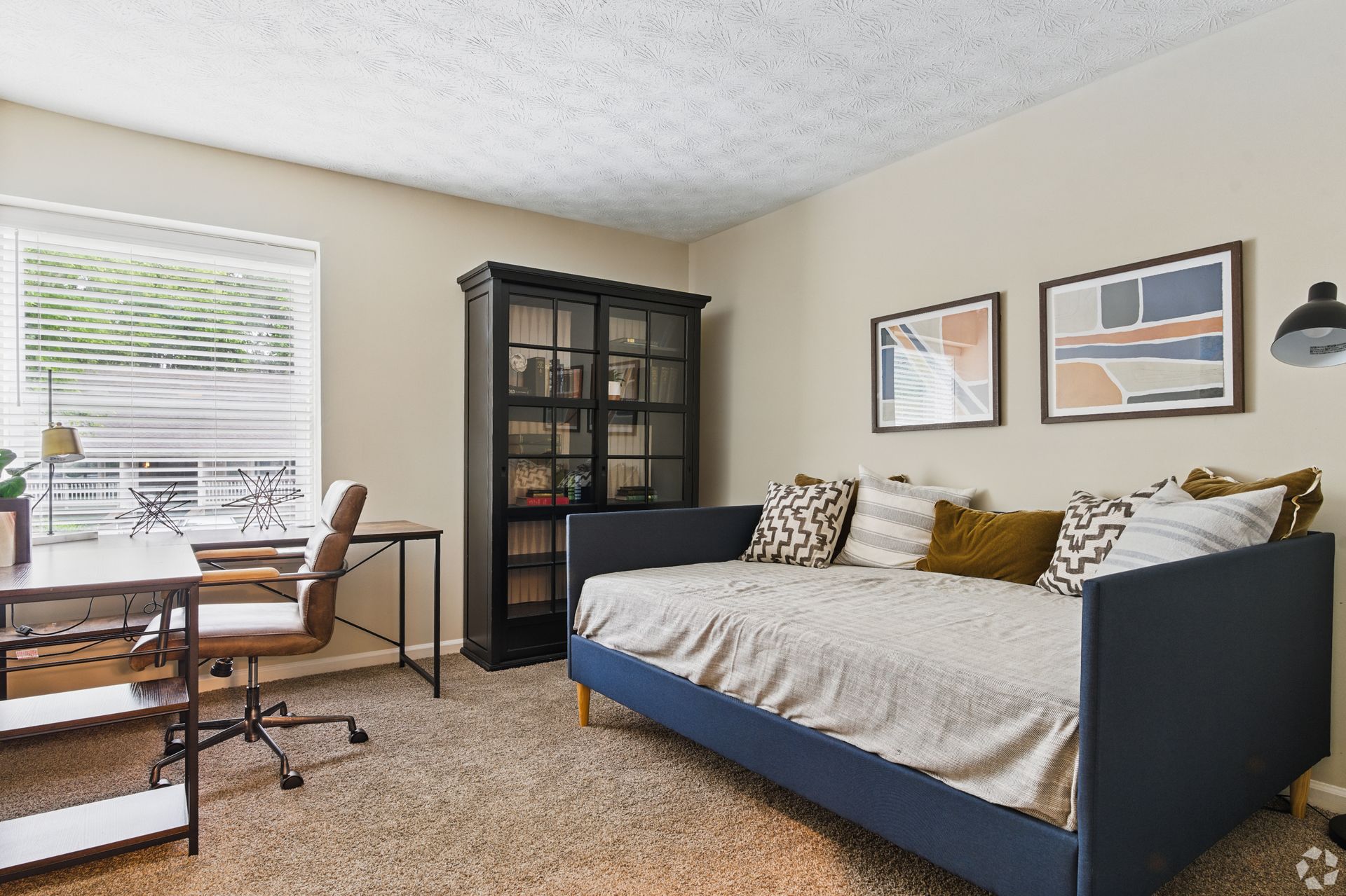 Cozy home office with a daybed, desk, and bookcase. Beige walls, neutral carpet, and blue accents.