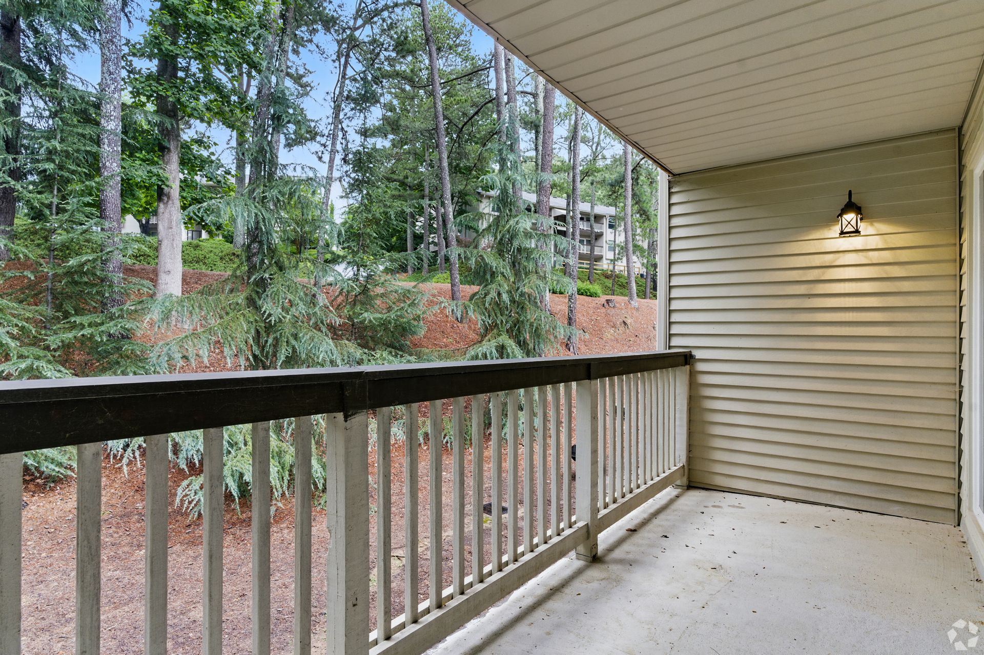 Balcony with white railing, forest view, beige siding, overhead light.