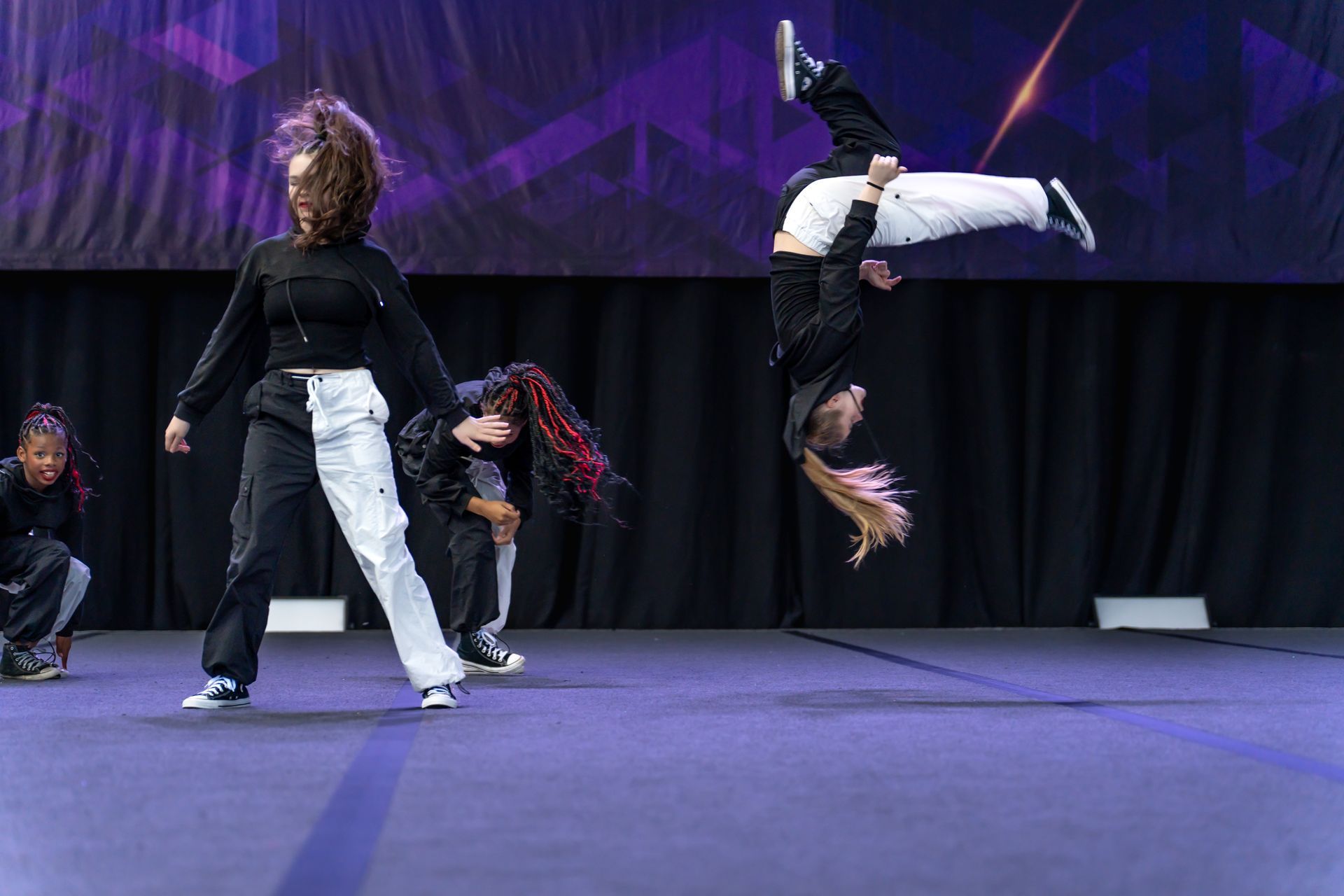 Dancers in black and white attire perform on a dark stage, with one mid-air in a flip.