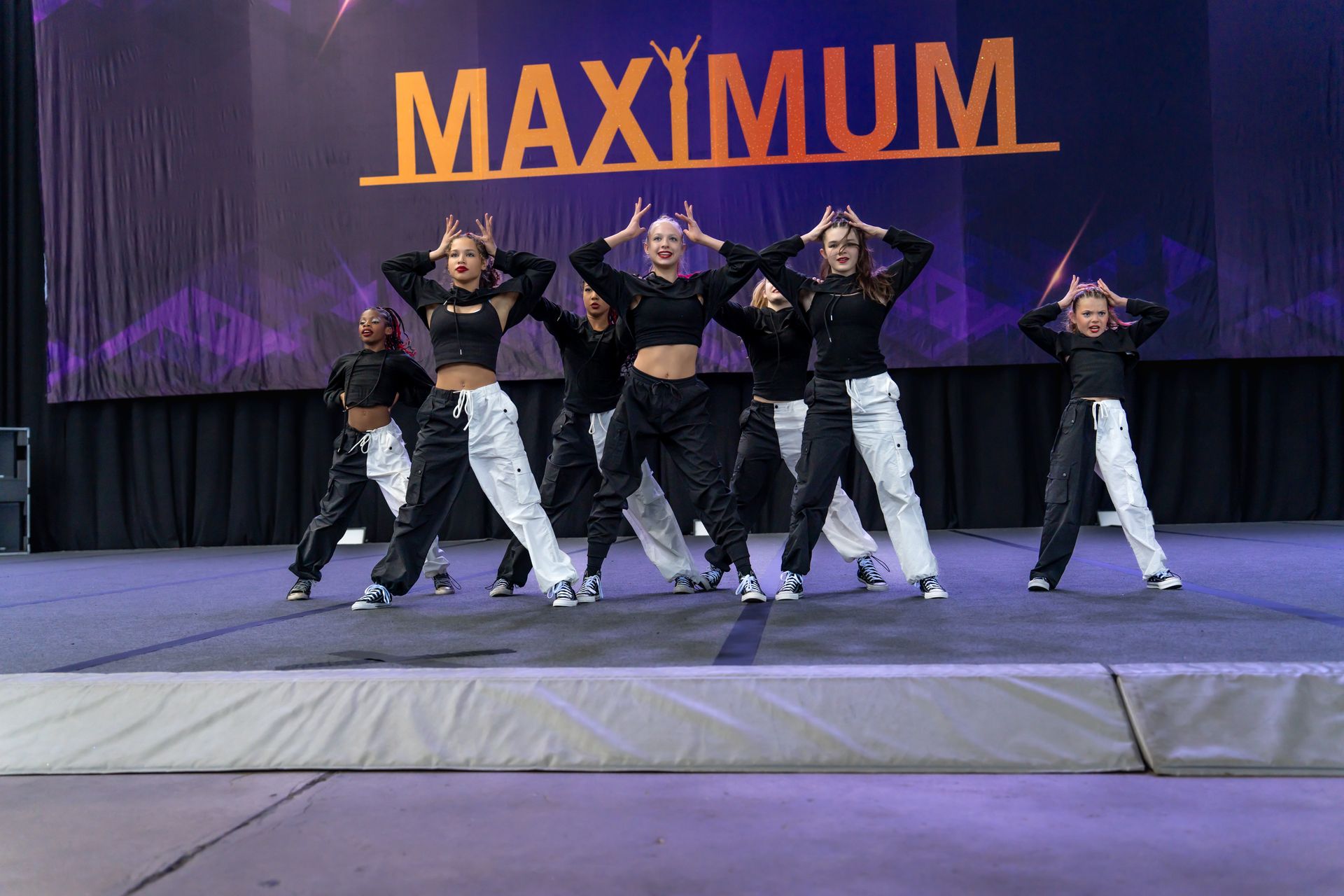 Five dancers in black and white outfits pose on a stage in front of a banner that reads
