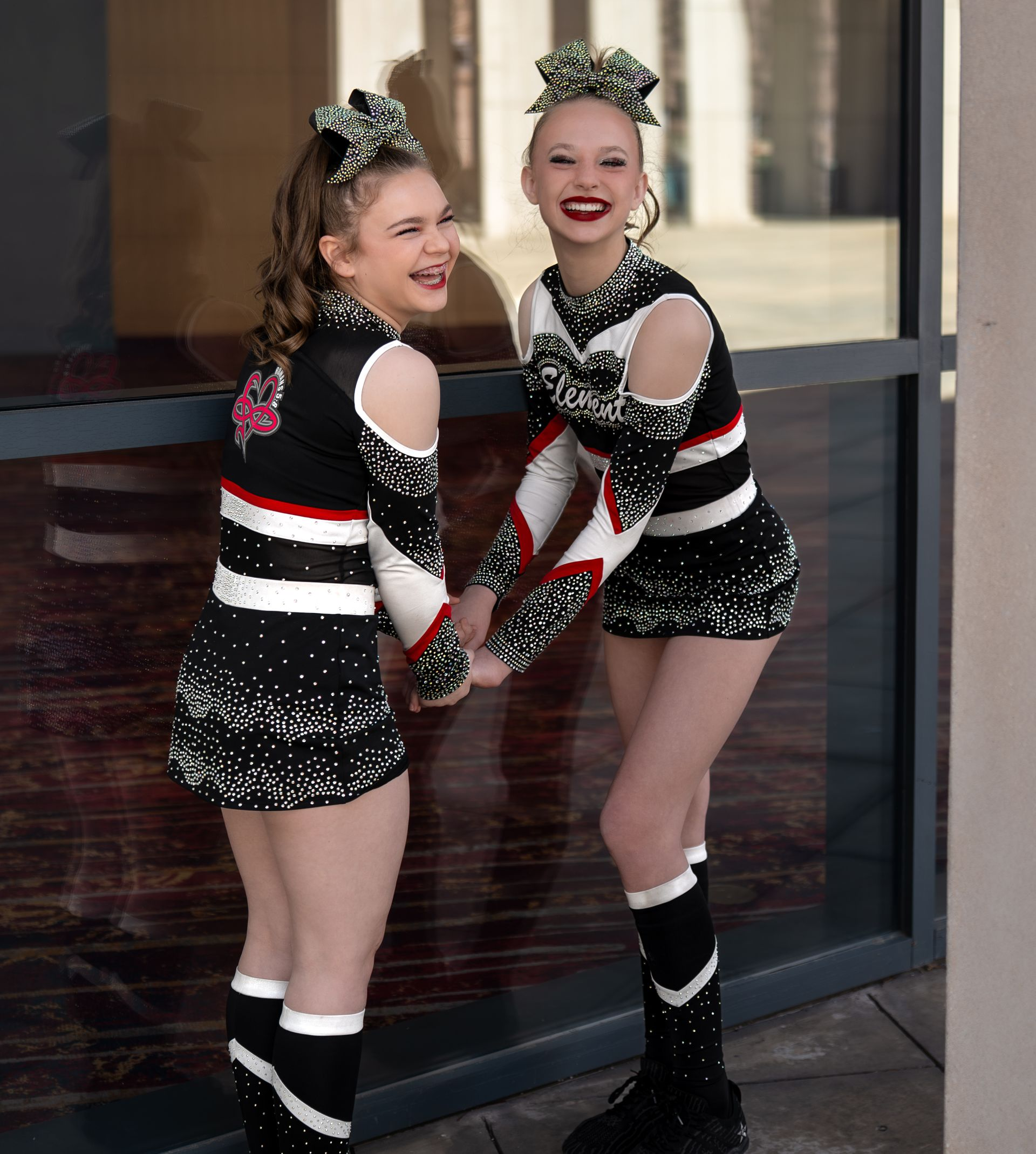 Two people in matching black, white, and red cheerleading uniforms and hair bows, laughing while holding hands outdoors.