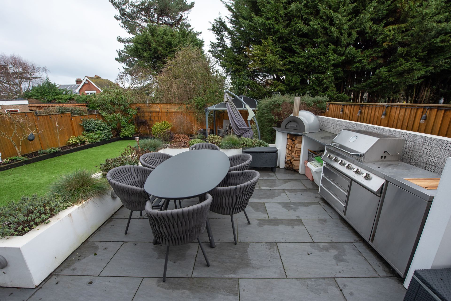 An outdoor kitchen with a dining table, chairs, grill, firewood stack, and garden with trees.