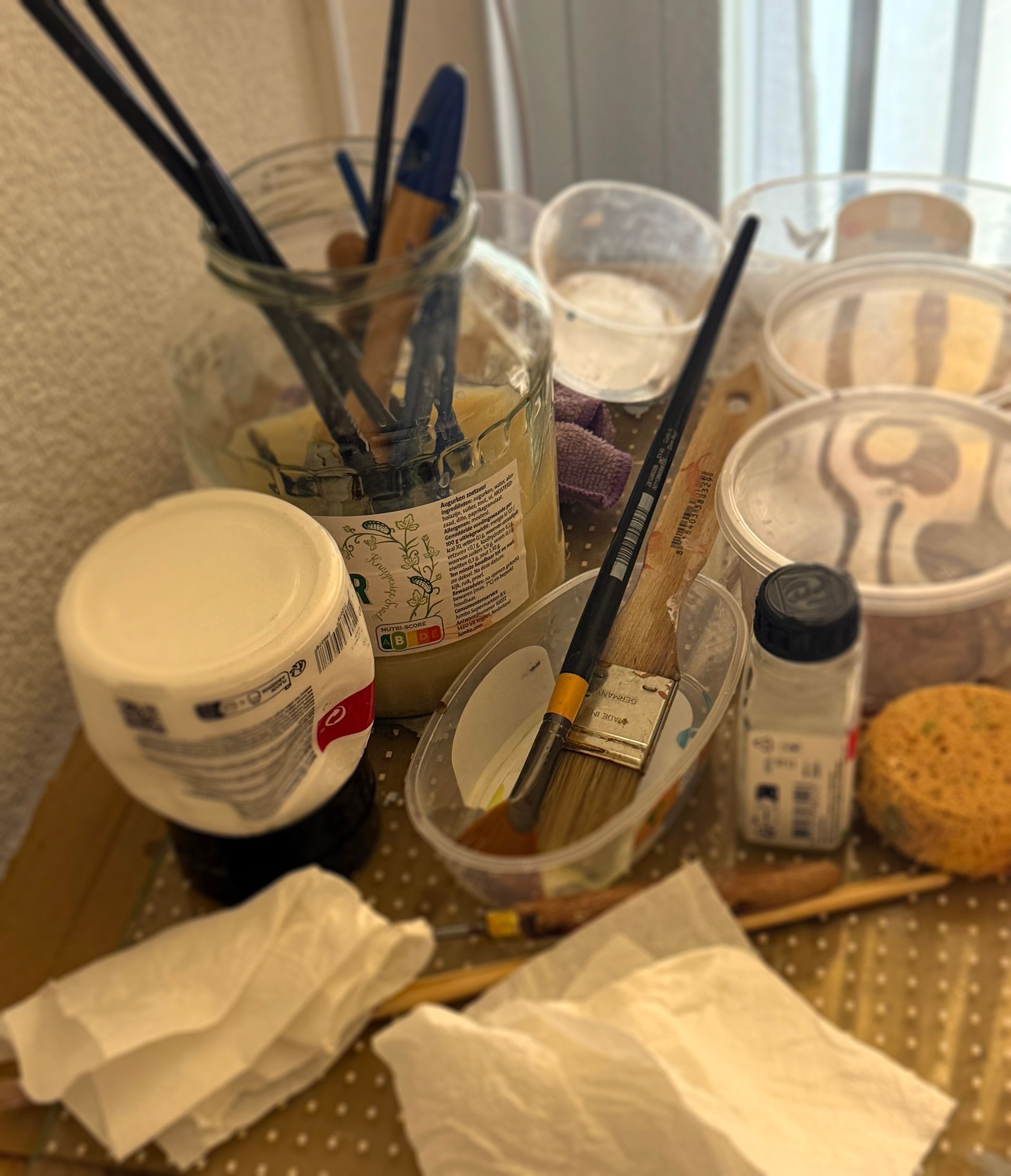 Een rommelige kaptafel met make-upkwasten, lotionpotjes, bekertjes, tissues en een lippenstifttube op een volgepakt aanrecht.