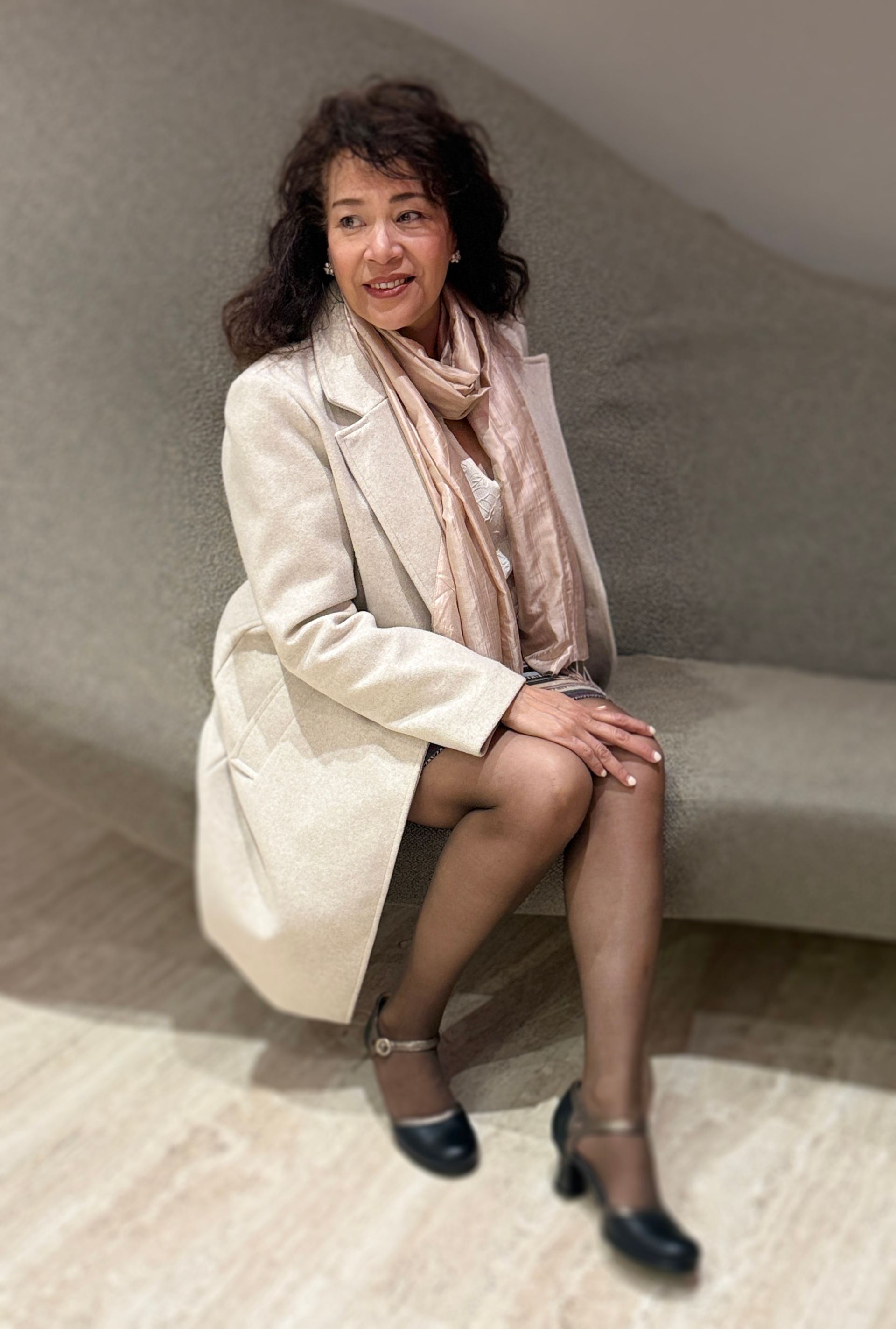 Vrouw zit op een grijze bank, gekleed in een beige jas en sjaal met zwarte hakken, en lacht naar de camera.