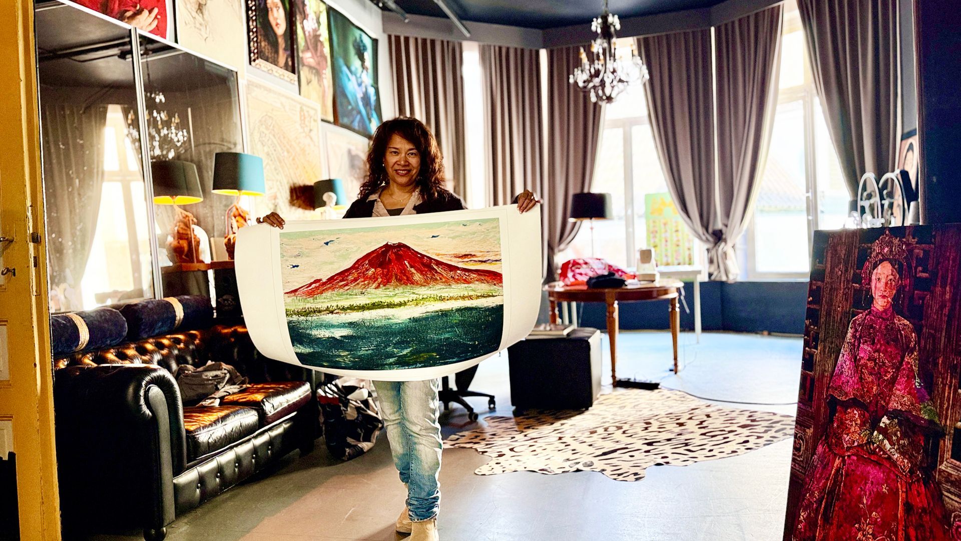 Vrouw met een schilderij van een rode berg en groen water in een lichte kunststudio.