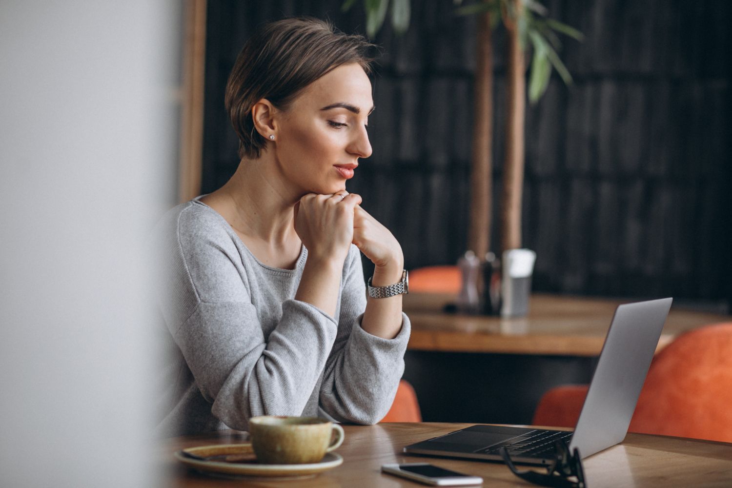 Woman sitting at a café table with a laptop, hands clasped, looking at the screen
