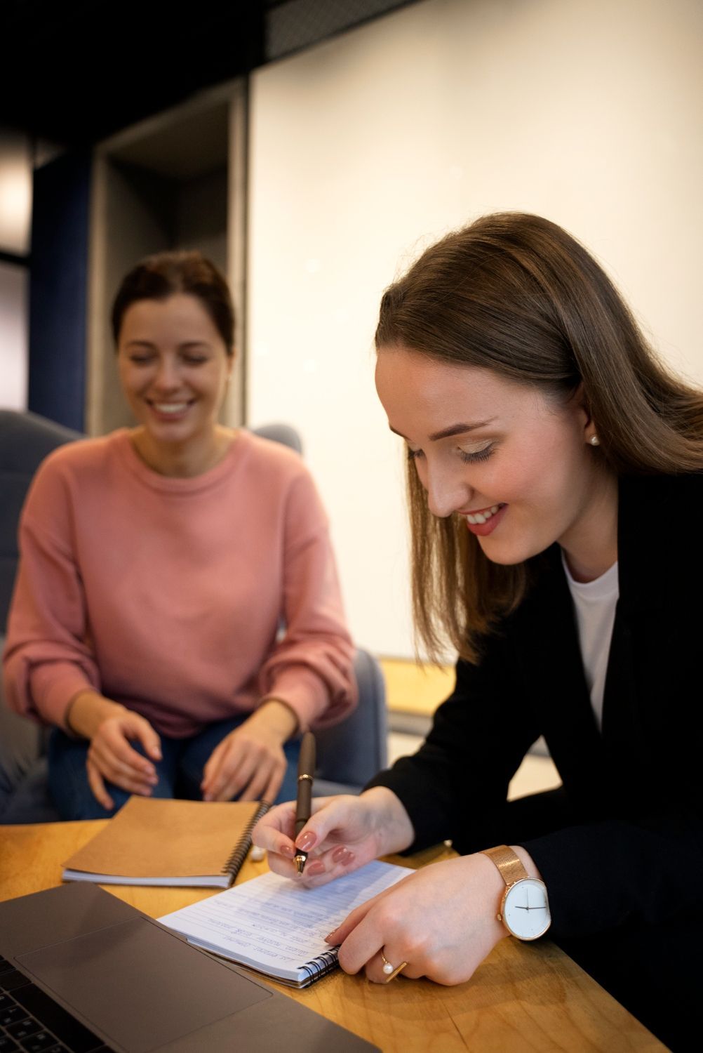 Two women in a meeting, one writing in a notebook while the other smiles at a table.