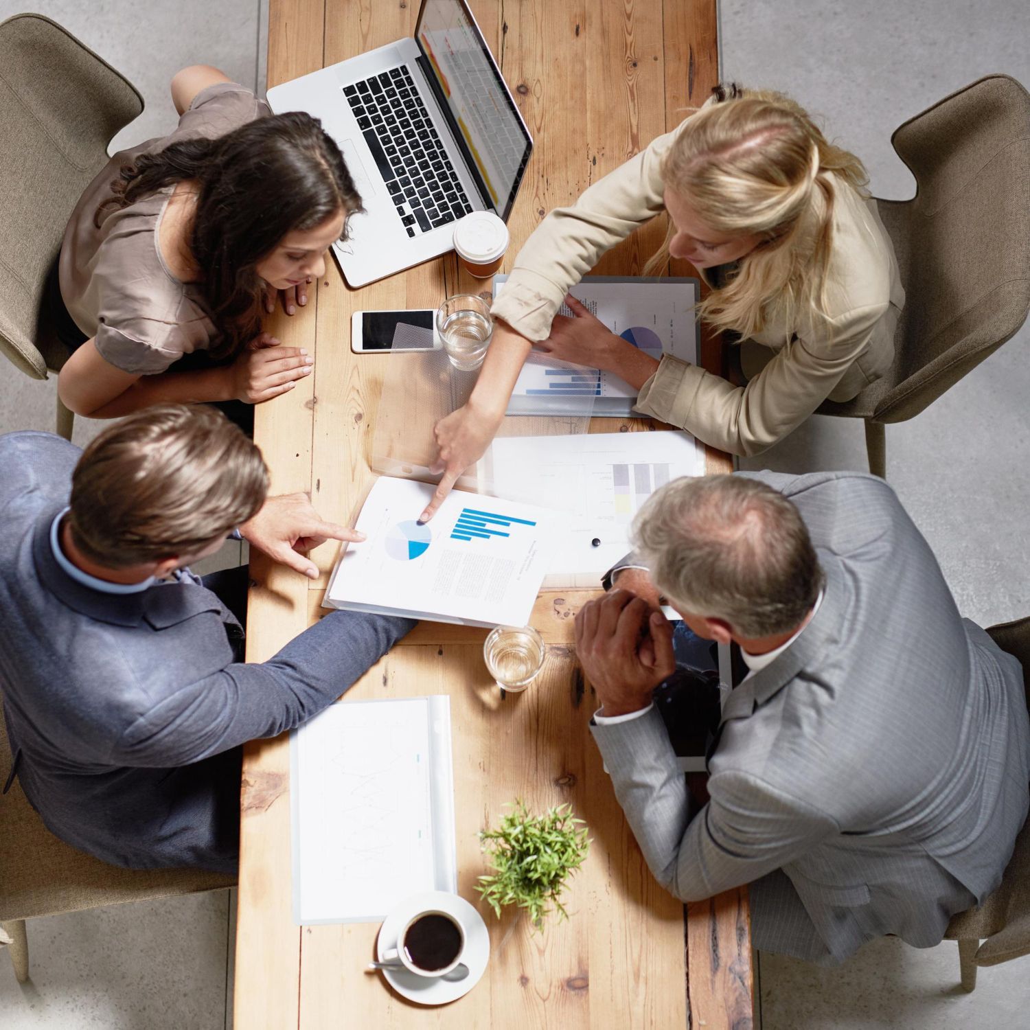 Overhead view of five people collaborating around a wooden table with papers, a laptop, and coffee.