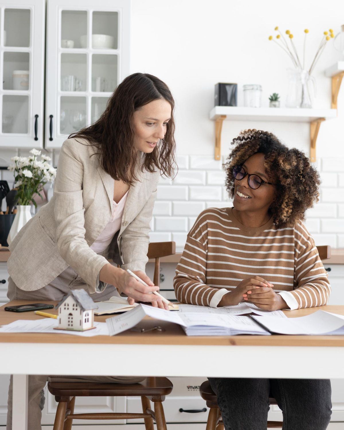 Two women reviewing documents at a desk in a bright office, one standing and pointing, one seated and smiling