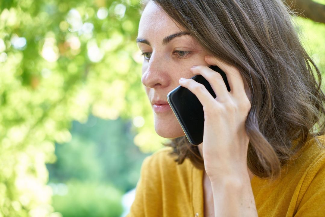 Woman in a mustard sweater talking on a smartphone outdoors with blurred greenery in the background