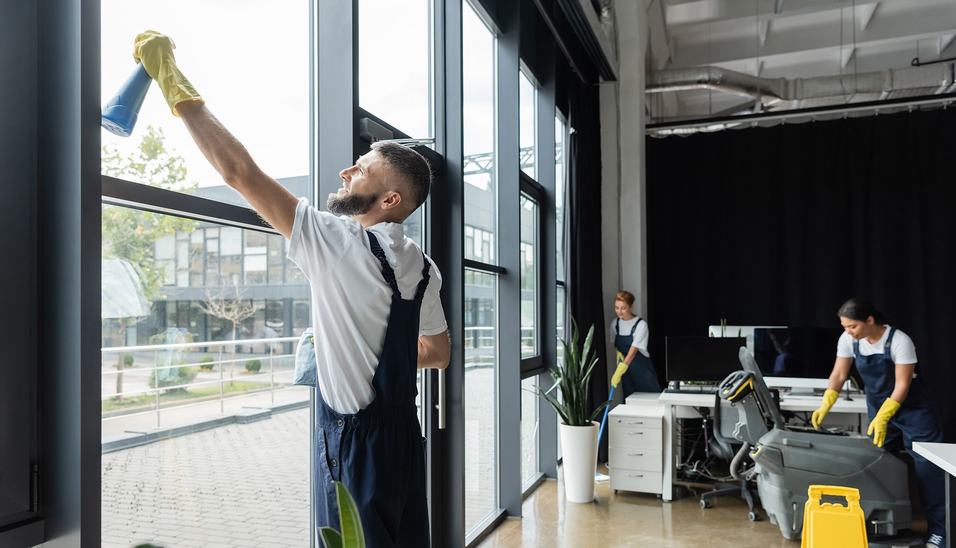 Person cleaning a large window in an office. Two people in the background clean desks and floor.