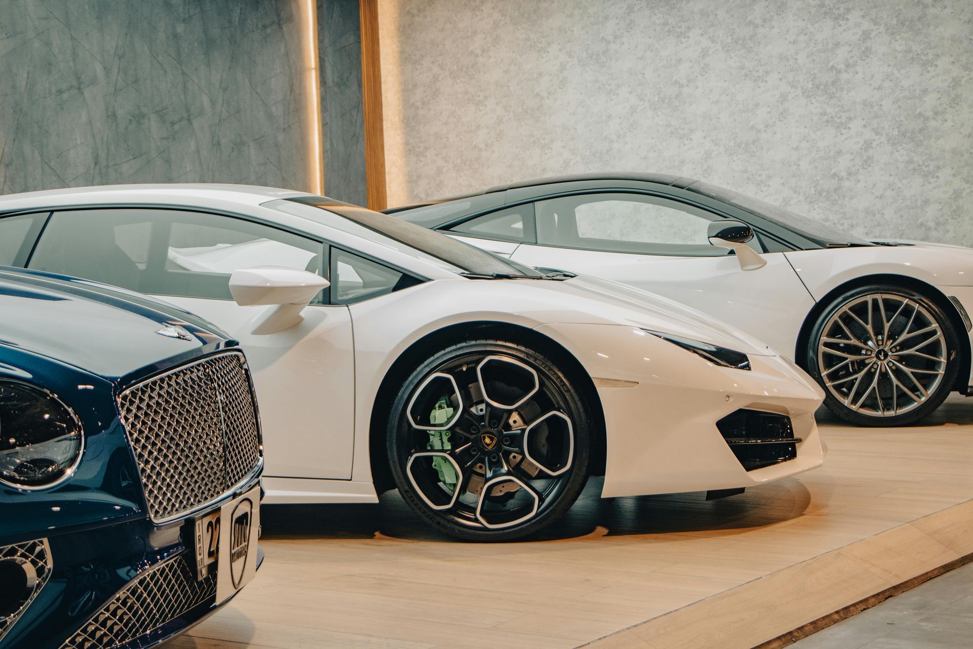 Three luxury sports cars in a showroom, including a white coupe in the foreground and a dark blue car on the left