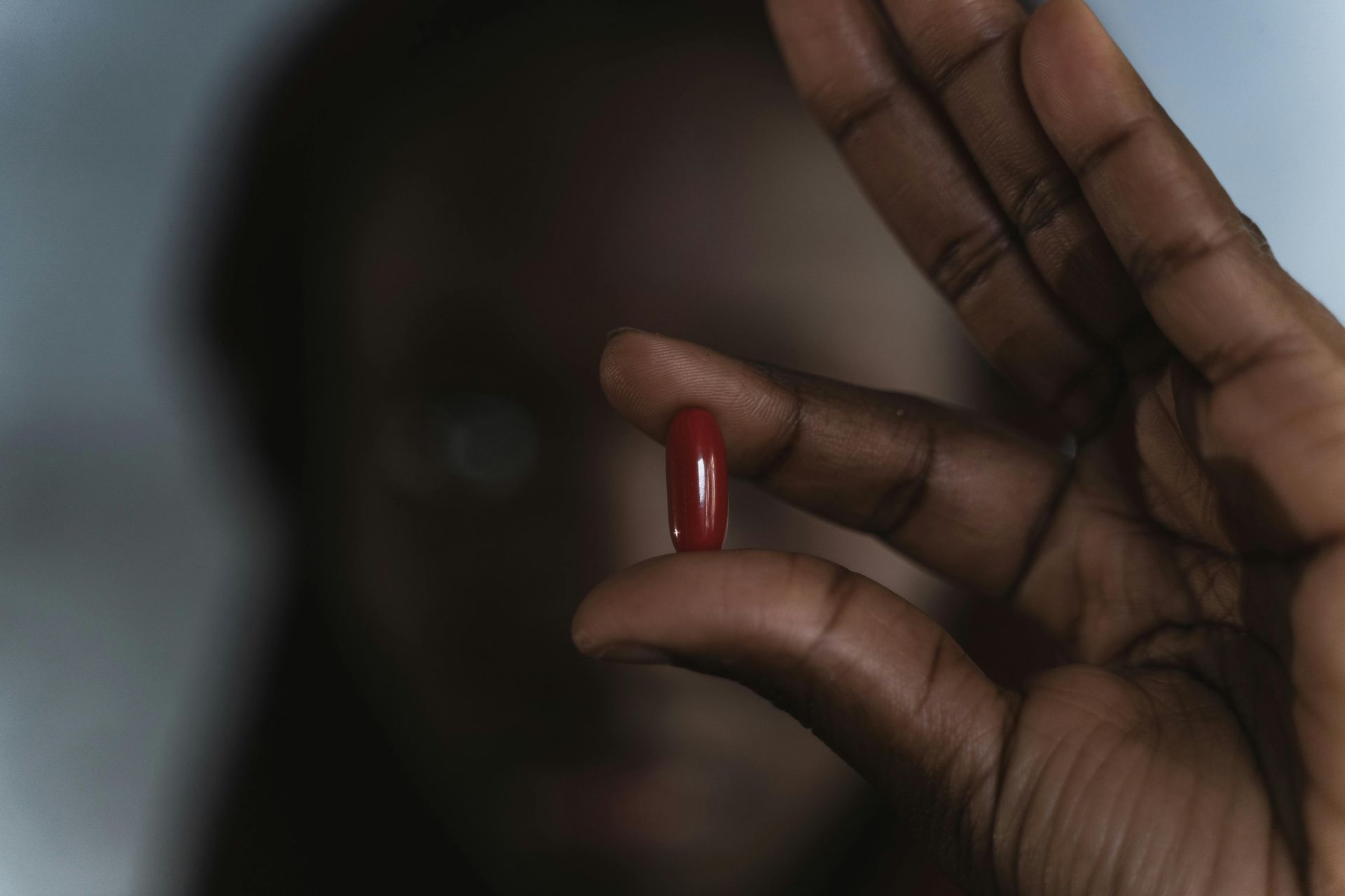 A hand holding a red capsule pill close to the camera, with a blurred face in the background.