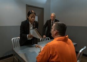 Two people question a seated man in an orange jumpsuit at a table in an interview room.