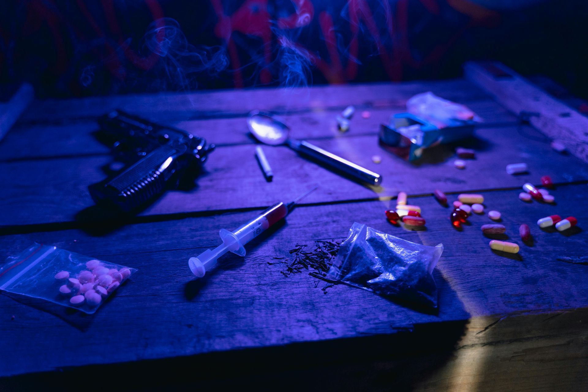 Scattered pills and a handgun on a wooden table under blue-purple lighting
