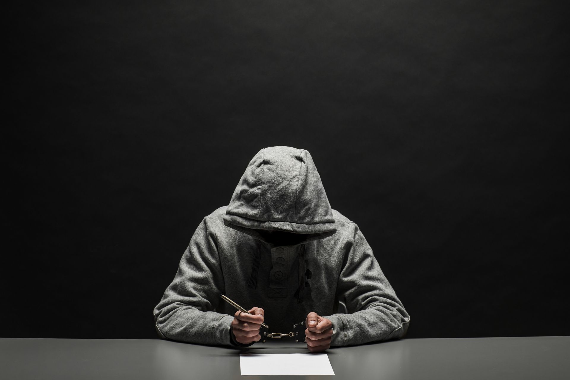 Person in a gray hoodie sits at a table, hands clasped, facing a blank sheet of paper in a dark room