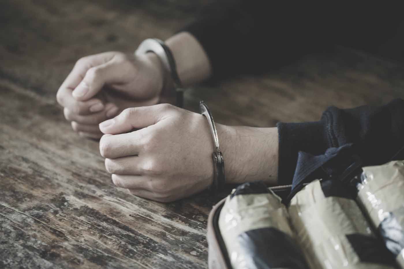 Handcuffed hands resting on a wooden table beside a wrapped bundle