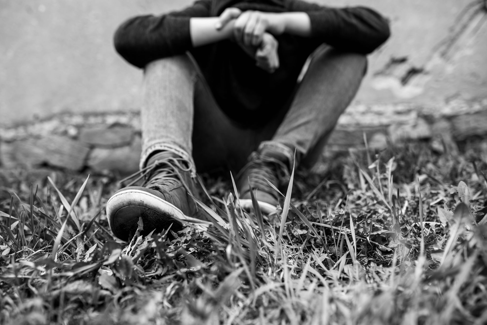 Black-and-white photo of a seated person with knees up, head down, in grass against a wall