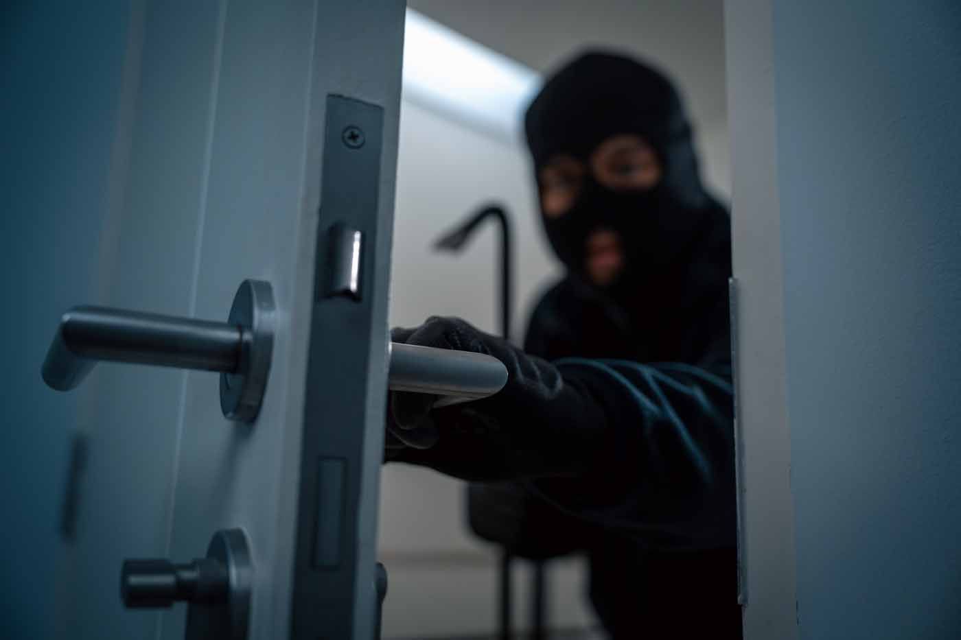 Masked intruder forcing a door open with a tool in a dim hallway