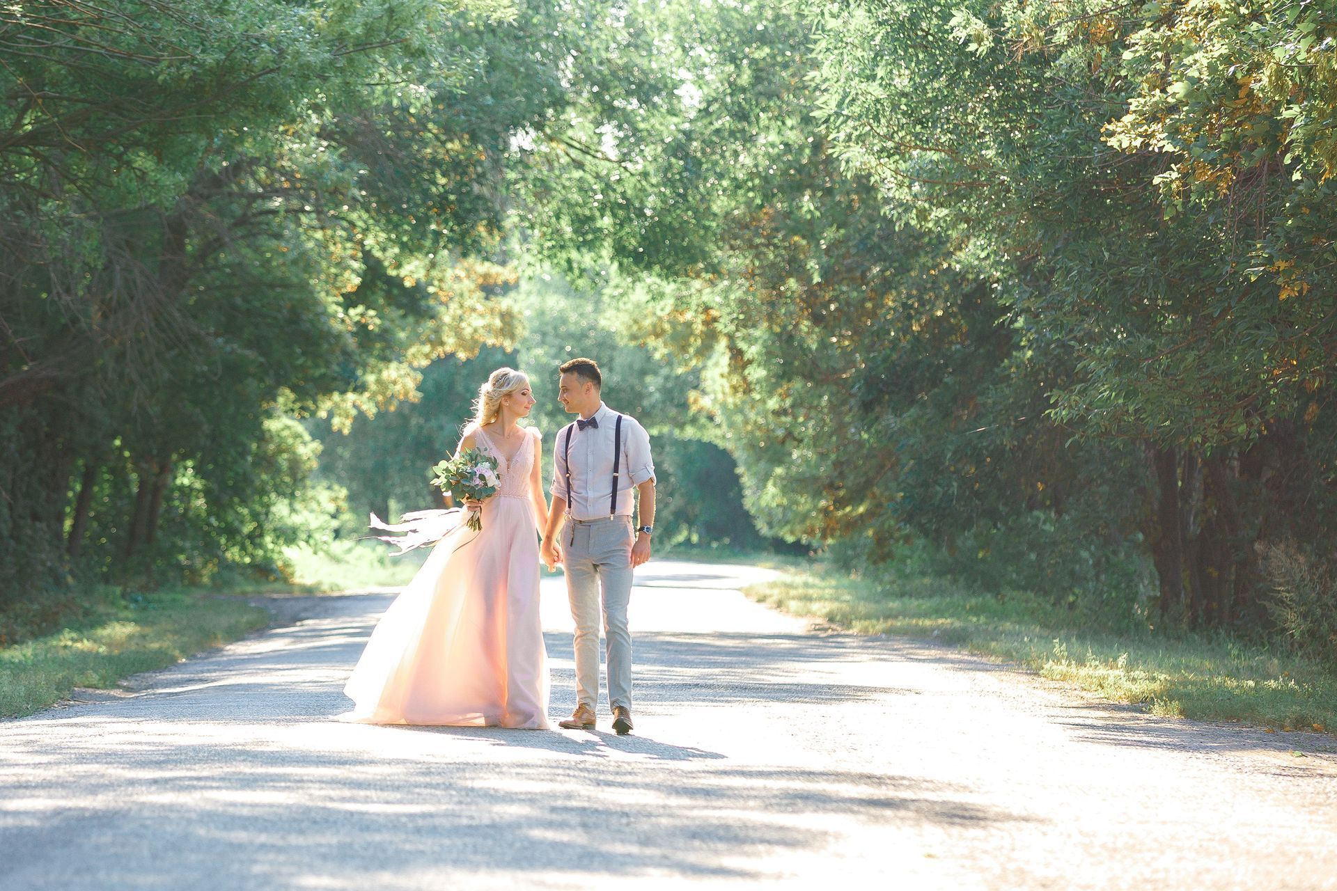 Couple walking hand in hand on a sunlit tree-lined road, bride in pink gown and groom in suspenders.