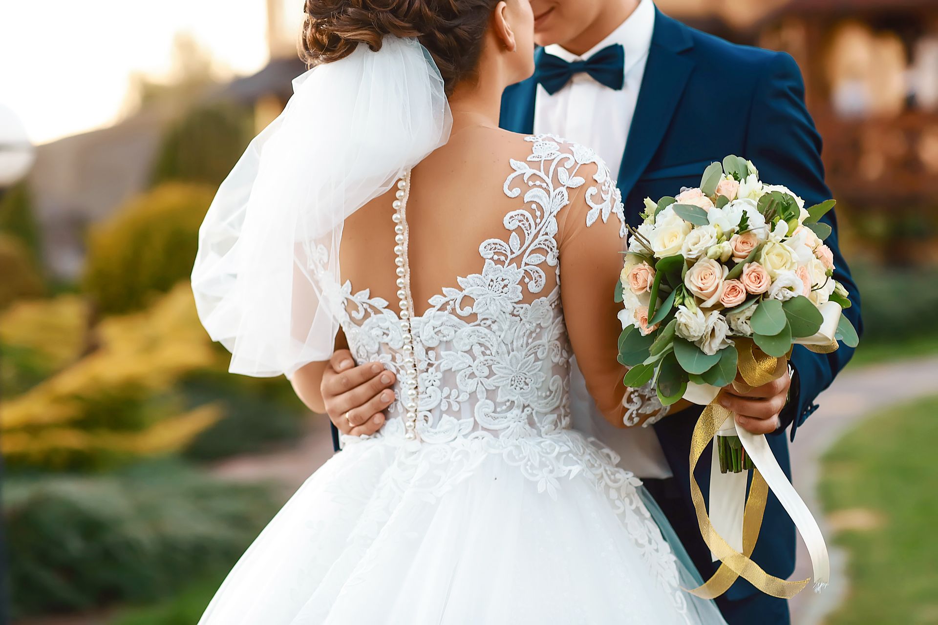 Bride and groom embracing outdoors, bride in white lace dress holding pastel bouquet.