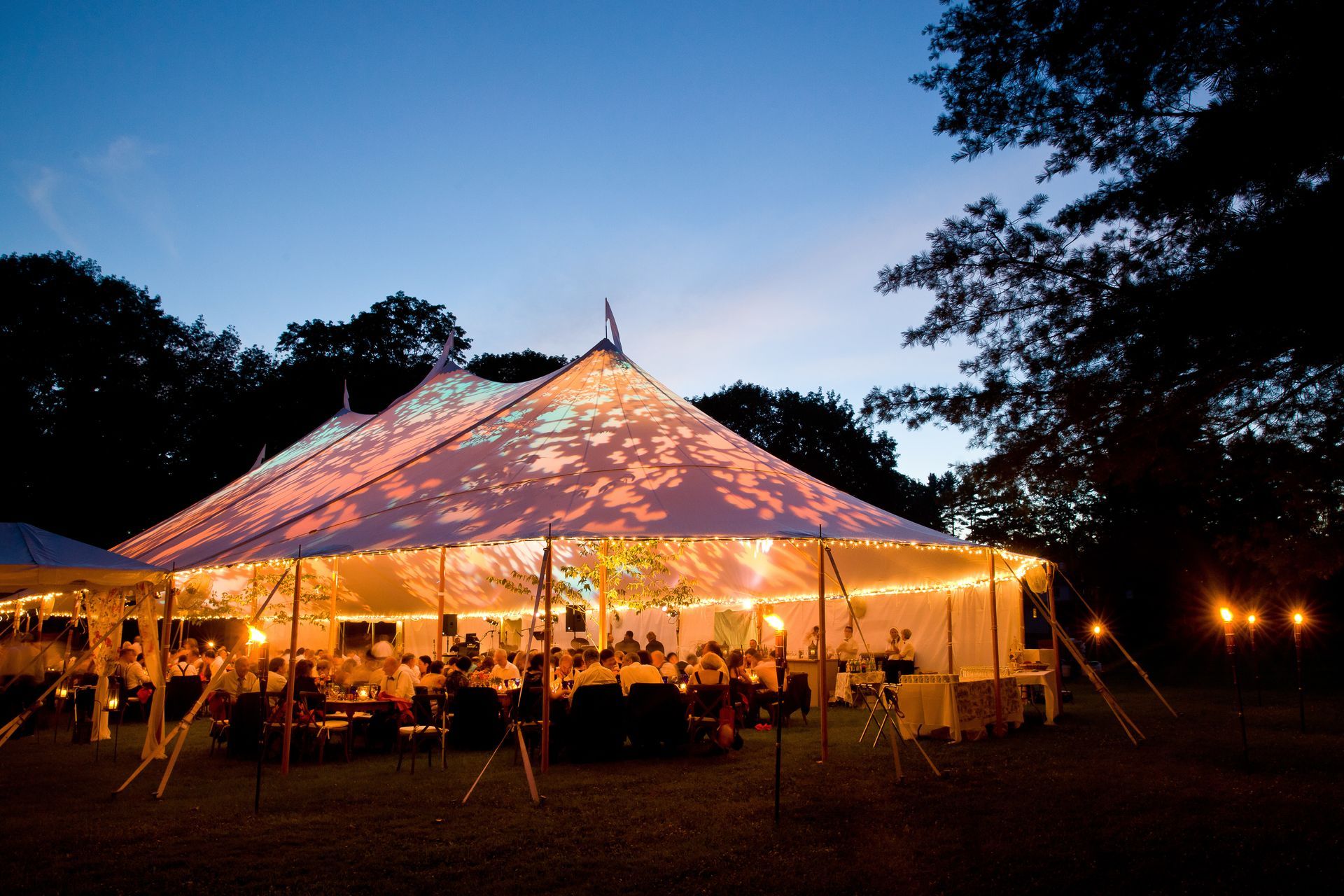 Lit outdoor event under a glowing white tent at dusk, with tables and warm lights in a dark field
