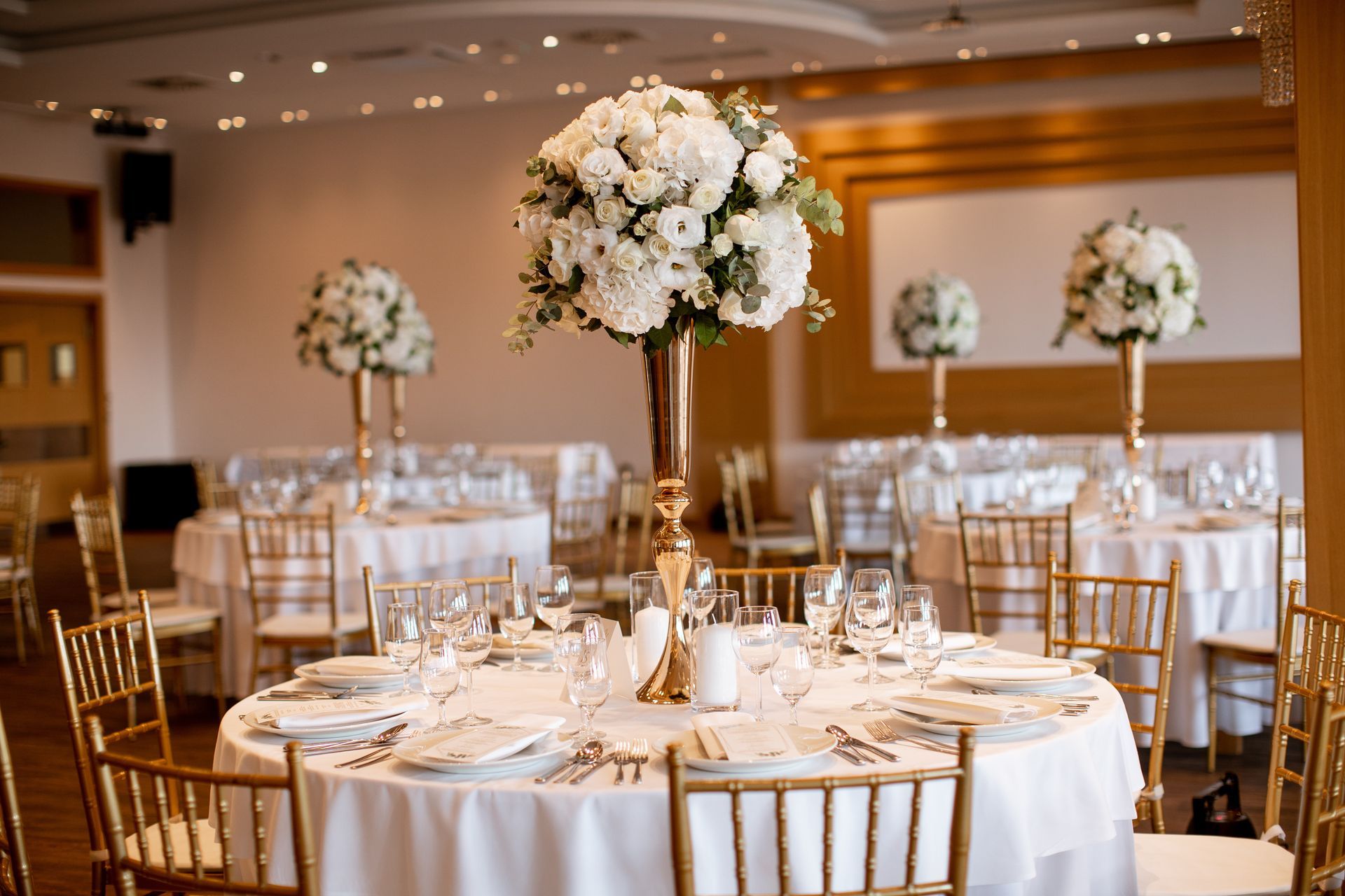 Elegant wedding reception hall with round tables, gold chairs, and tall white floral centerpieces.