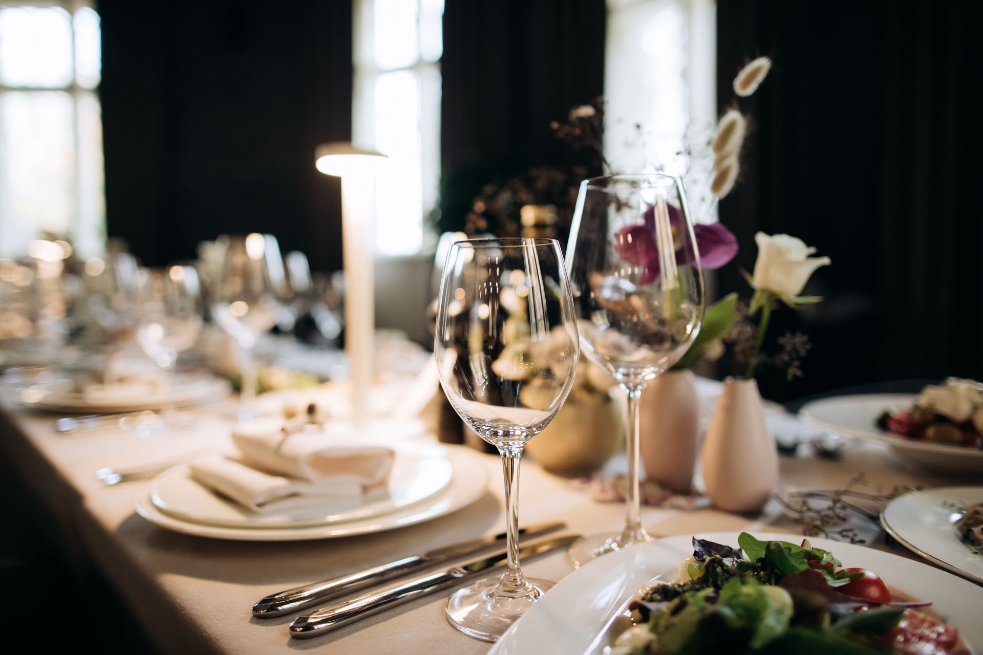 Elegant dining table set with wine glasses, white plates, silverware, and floral centerpieces in soft light