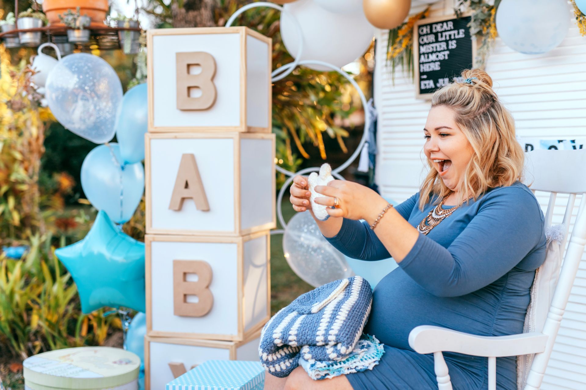 Woman seated at a baby shower, holding a clear balloon beside stacked “BABY” blocks and blue-white decorations