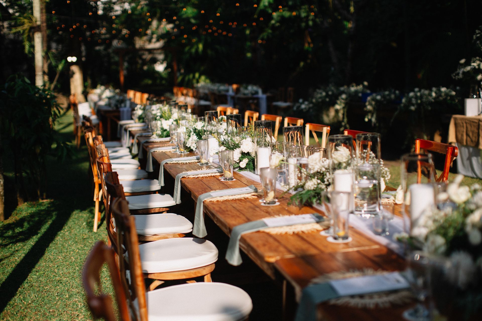 Long outdoor banquet table set with white chairs, glassware, and greenery in a garden setting