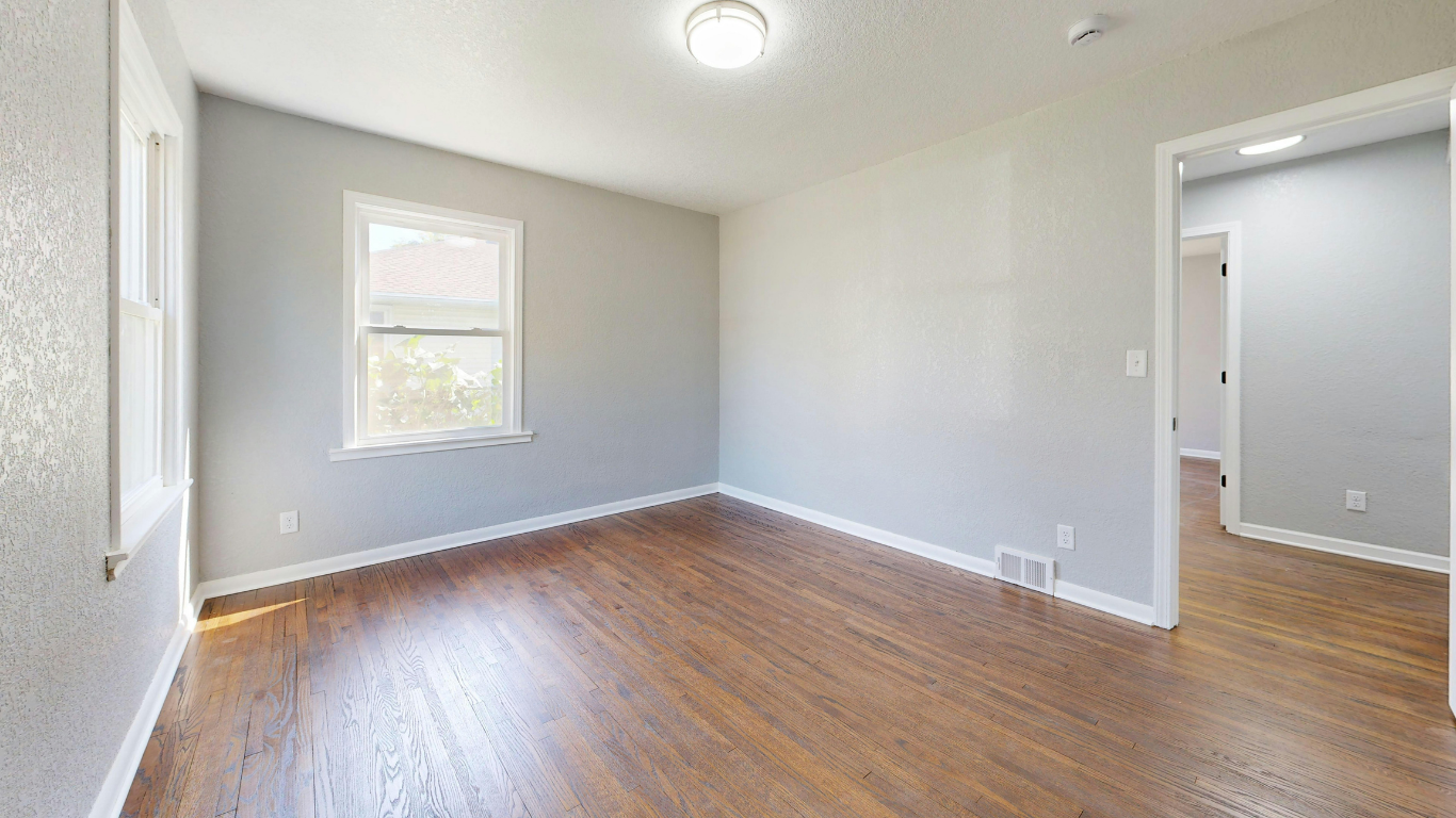 An empty room with light gray walls, brown hardwood floors, a single window, and a doorway to another room.