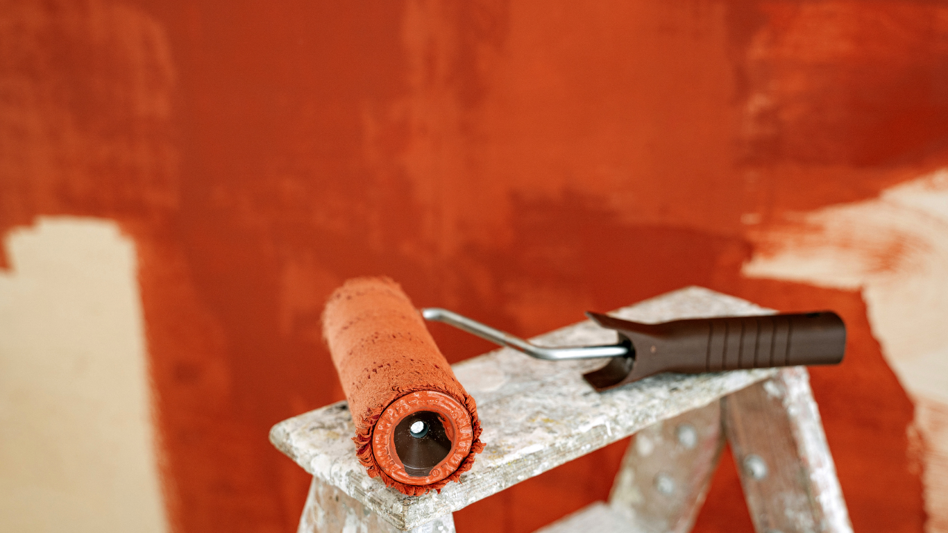 A paint roller covered in orange paint rests on a small metal step ladder against a wall partially painted orange.